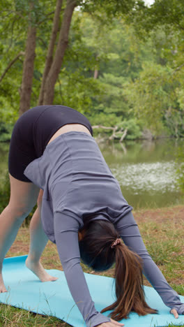 Vertical-Video-Of-Woman-Wearing-Sports-Clothing-Doing-Yoga-Stretches-On-Mat-In-Forest-By-Lake-Or-River-Enjoying-Peace-And-Beauty-Of-Nature-Shot-In-Real-Time-2